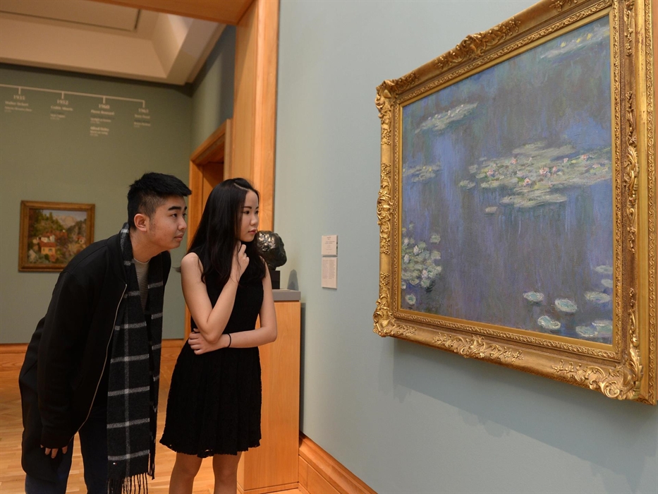 a young man and woman look at a painting. The painting is in a gold frame and shows blue water of a pond with waterlilies floating on the surface of t