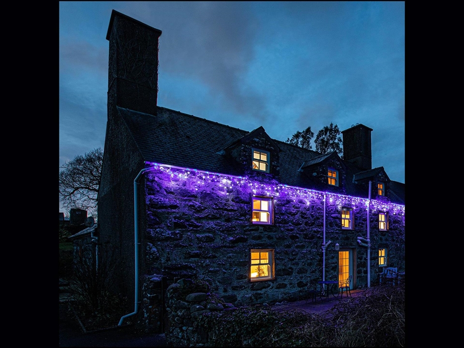 Betws Bach Farmhouse with Festive Lights hanging along the front. Blue icicles on at evening with a dark sky and lights on, inside the farmhouse