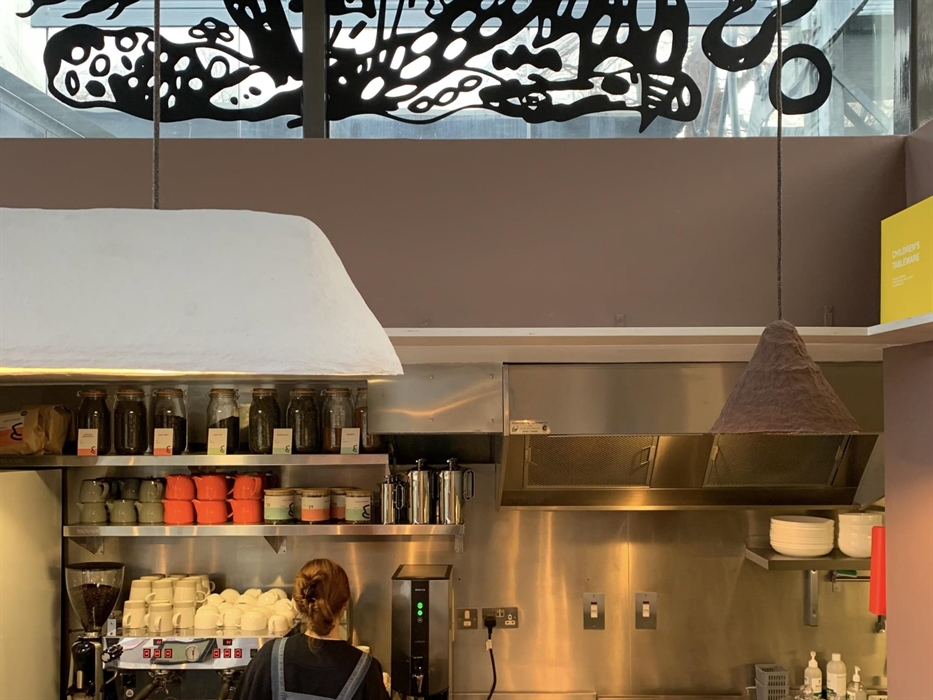 Cafe kitchen with shelves stacked with cups, teapots and jars. a woman with her back to us is operating the coffee machine.