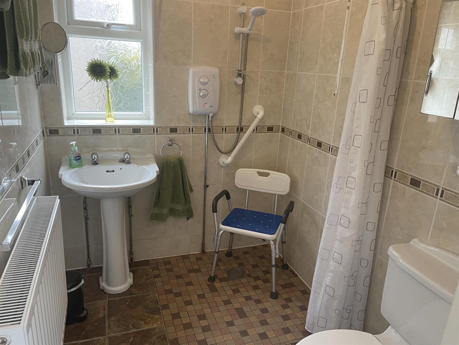 shows sink, window, electric shower, radiator, shaving mirror, electric shaver socket, mirror cupboard, hand grip and a height adjustable shower chair