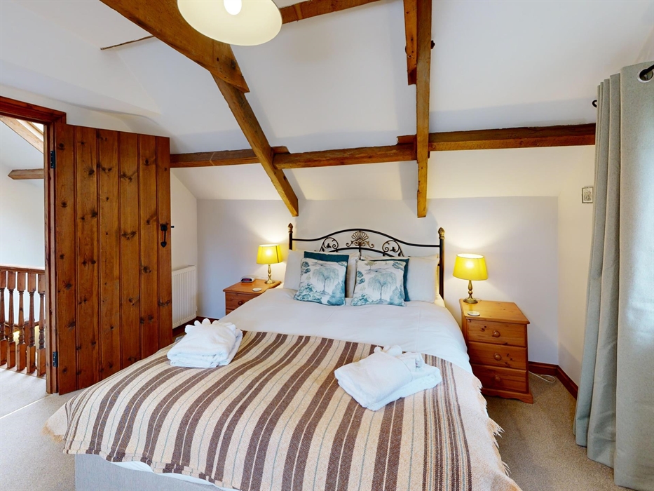 A bright bedroom with exposed wooden beams, double bed, wooden wardrobe, and soft lighting.