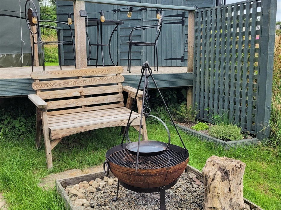 In front of the deck at the yurt, wood bench with wood framed gravel square containing Kadai fire pit with grill and cast iron skillet on the tripod a