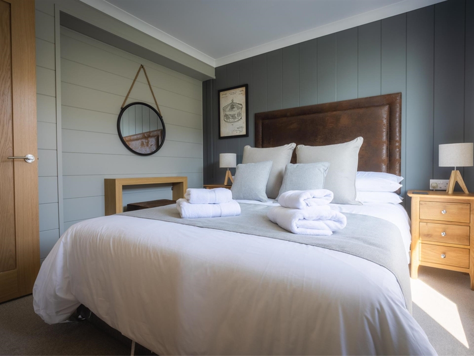 Double bed with brown, faux leather headboard, bedside table with lamp, Carousel wall print and round mirror hanging from rope above dressing table.