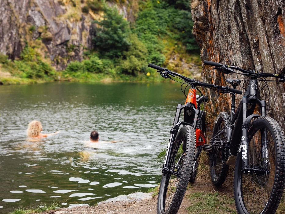 Two people swim in a lake with their bikes resting in the foreground.