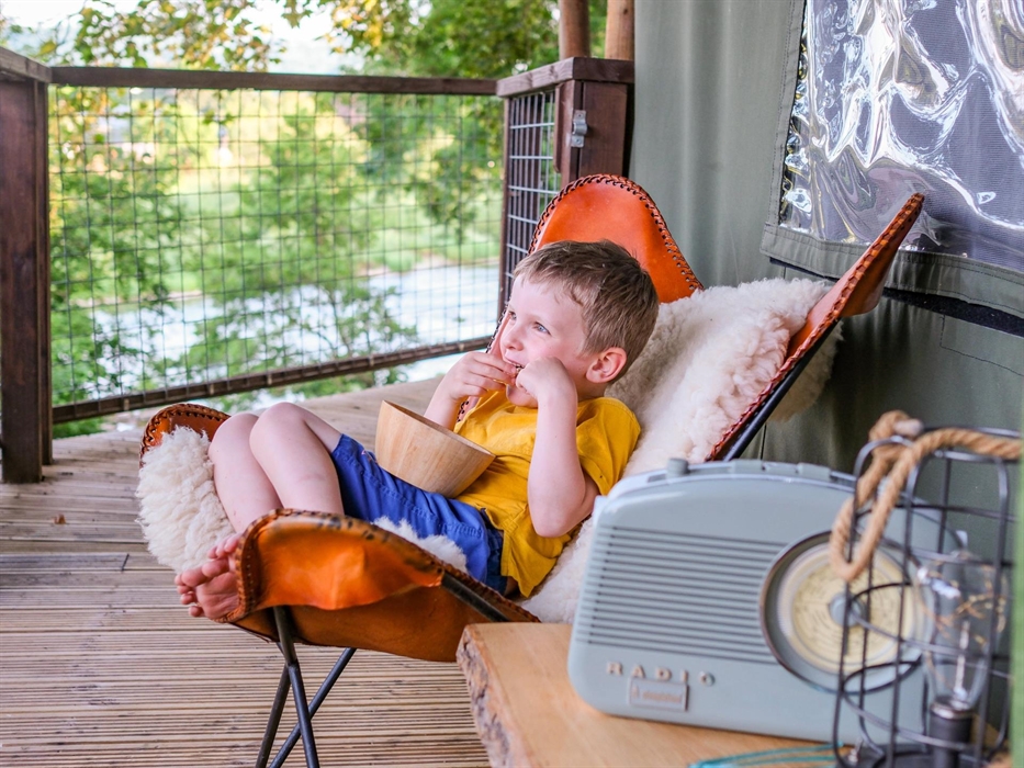 A child sat in a cozy chair on the decking of a safari tent eating popcorn watching the river.