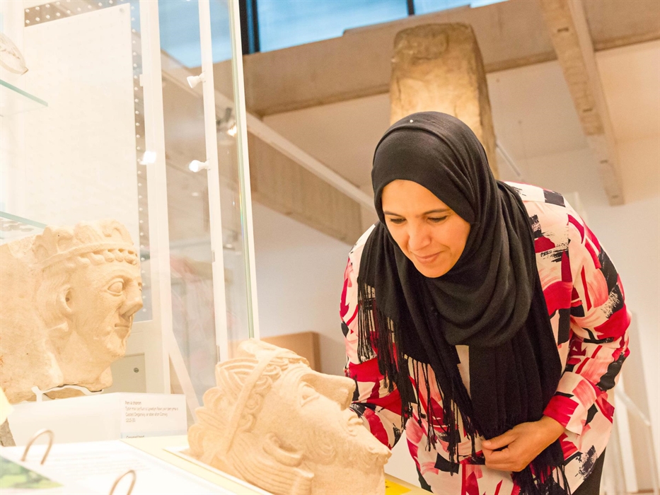 a lady wearing a black hijab and red patterned blouse is leaning forward to look at a stone carving. The carving shows the head of a medieval prince.
