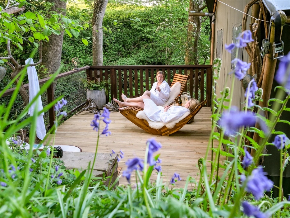 Friends relax on the deck of a safari tent in the woods.  Glamping Holidays in Wales.  Bluebells