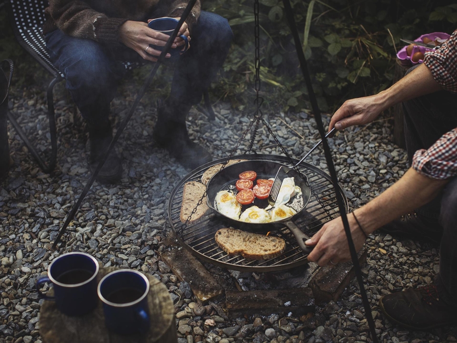 A campfire scene with people sitting around it (just visible). Cooking on a barbecue over the fire is some toast, frying eggs in a pan and fried tomat