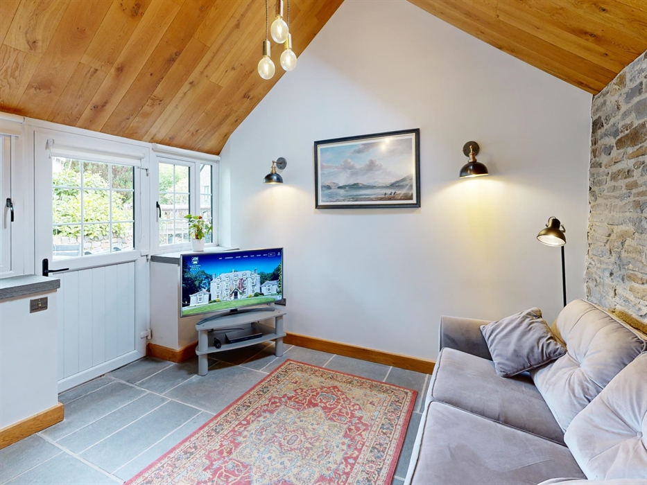 A comfortable living room with a grey sofa, exposed stone wall, flat-screen TV, and a large window and door overlooking a garden. The space features a