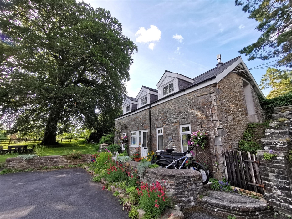 A characterful stone-built cottage with white-framed windows, surrounded by colourful flowers and climbing plants. A bike rests by a bench in the fron