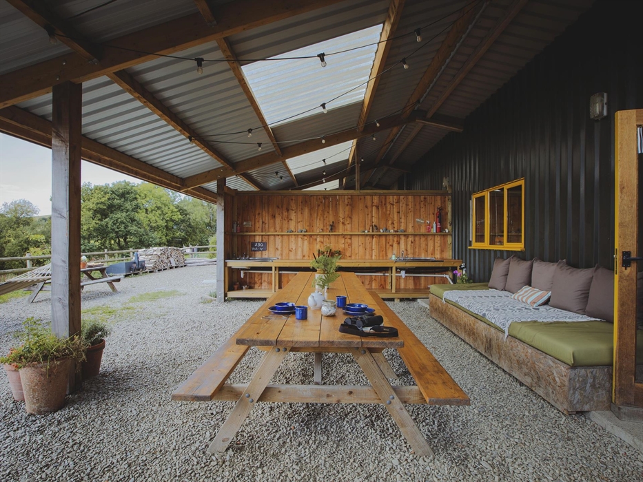 A large picnic table sits centre of an outdoor covered living space. There is a sofa to the right and washing up sinks in the background. Festoon ligh