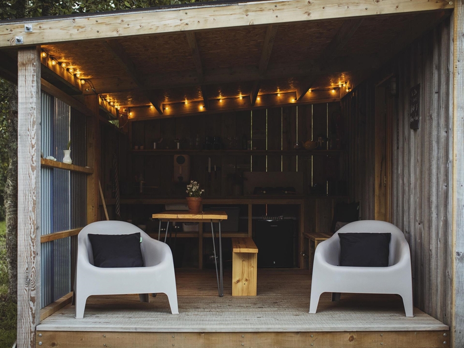 A covered camp kitchen with festoon lighting above. A dining table and two lounging chairs in the foreground.