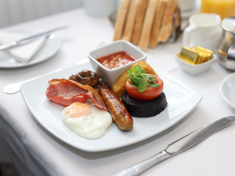 A plated breakfast with a fried egg, bacon, sausage, grilled tomato, hash brown, pan fried mushrooms, baked beans in a ramekin and black pudding.
