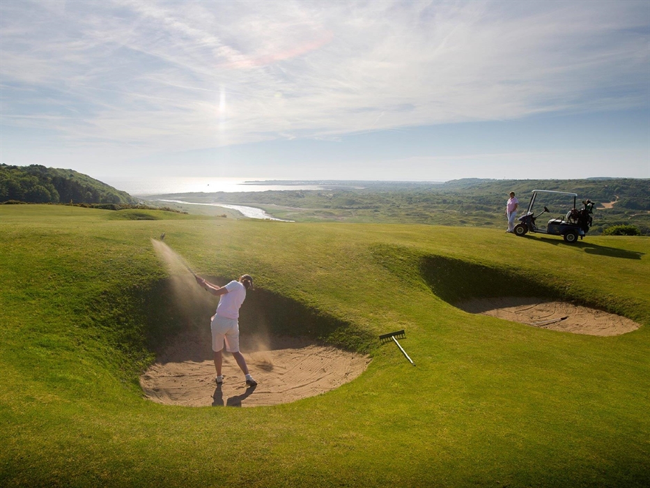 A golfer chipping out of a sand bunker at Southerndown golf club with a view of the coast behind.