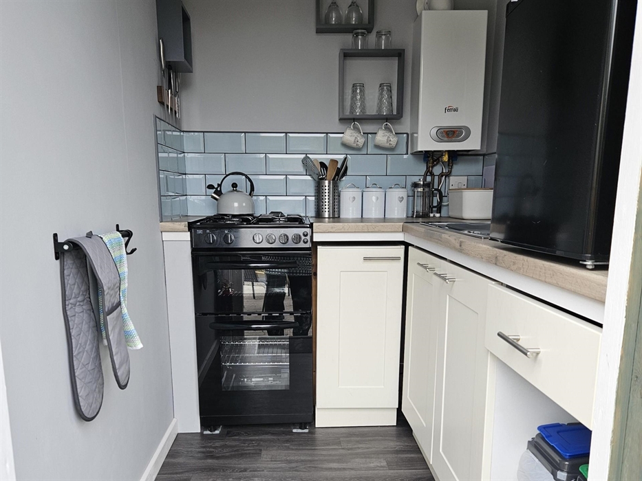 A gas stove with hob top kettle, boiler, sink and black counter top fridge. TUtensils, tea,coffee,sugar canisters, coffee pot are on the work top. The