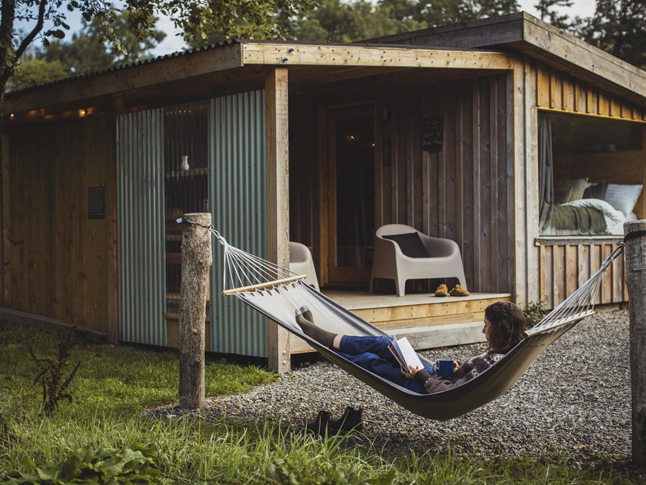 A woman lies in a hammock whilst reading a book. In the background is a wooden cabin with some lounge chairs on a deck and a large window with an unma