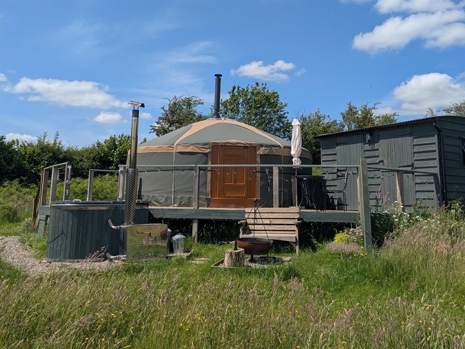 Green Yurt on a raised deck with dark green wood hut on right of photo, wood fired hot tub to left. Bench and fire pit to front surrounded by wild flo