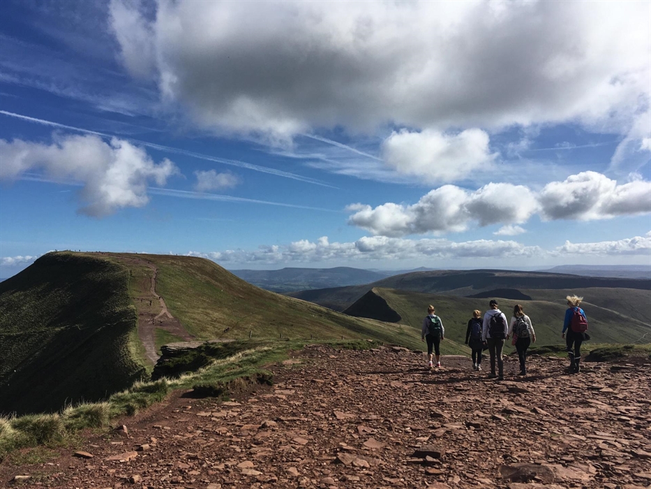 Five people walking into the distance on top of a ridge, facing a peak with blue skies and fluffy clouds