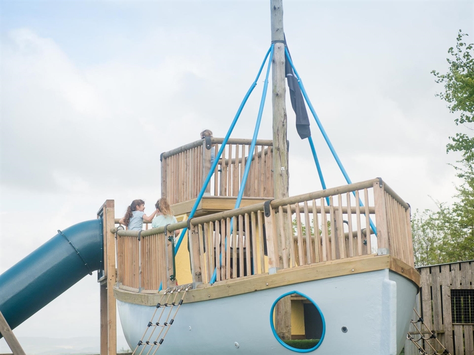 A wooden pirate ship rises from the play area, complete with lookout deck, rope bridge and portholes. Surrounded by green hills, it invites little buc