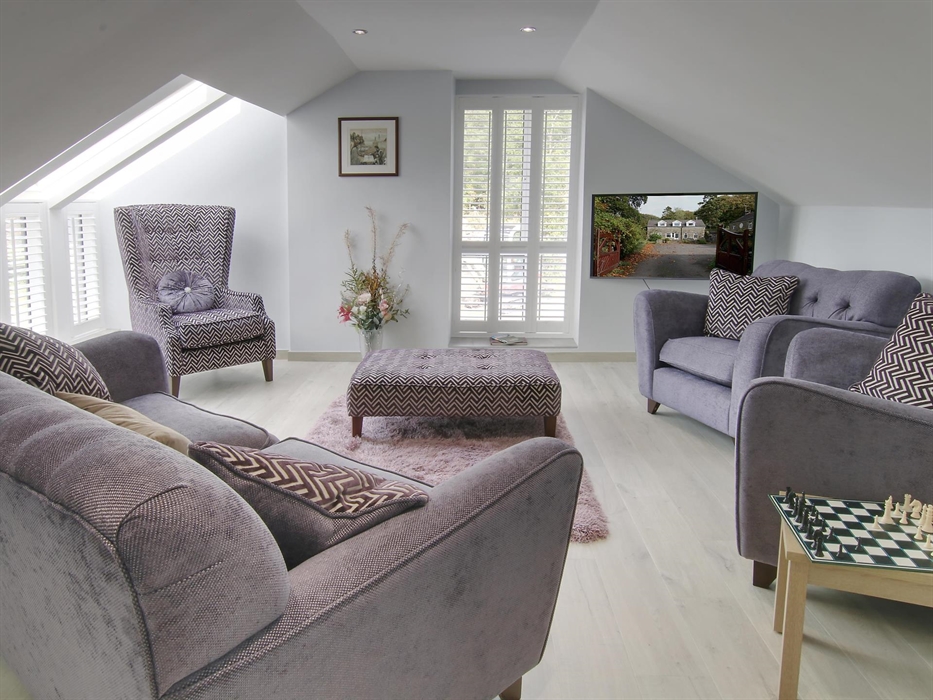 Elegant living room with grey armchairs, patterned cushions, and a chess table, bathed in natural light from skylights and tall windows.