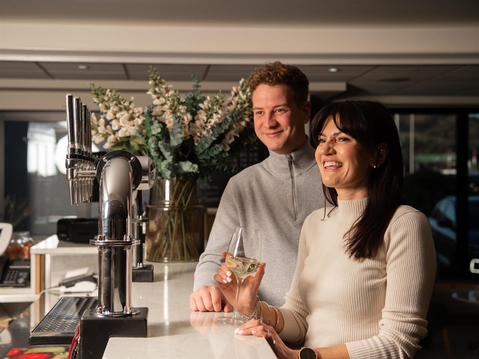 A woman and a man at a bar with the woman holding a glass of wine and a vase of flowers behind them