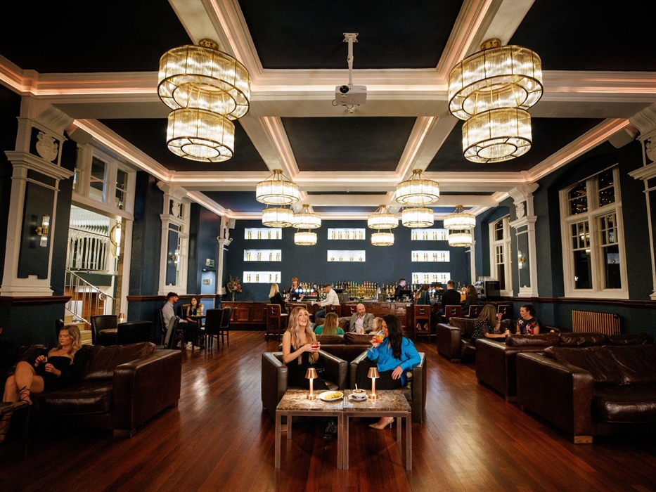 Image shows a large bar-lounge, with some guests sitting on sofas and drinking cocktails, whilst others sit at the bar at the rear.