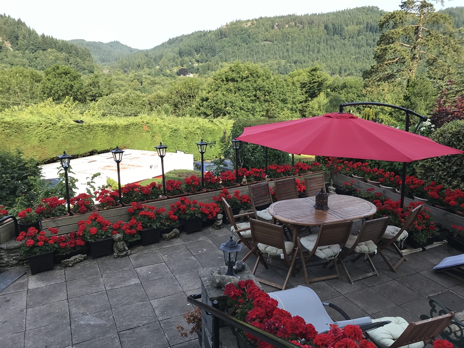 A stone‑paved patio with a round wooden table and six cushioned wooden chairs beneath a large red umbrella. The patio is lined with vibrant red flower