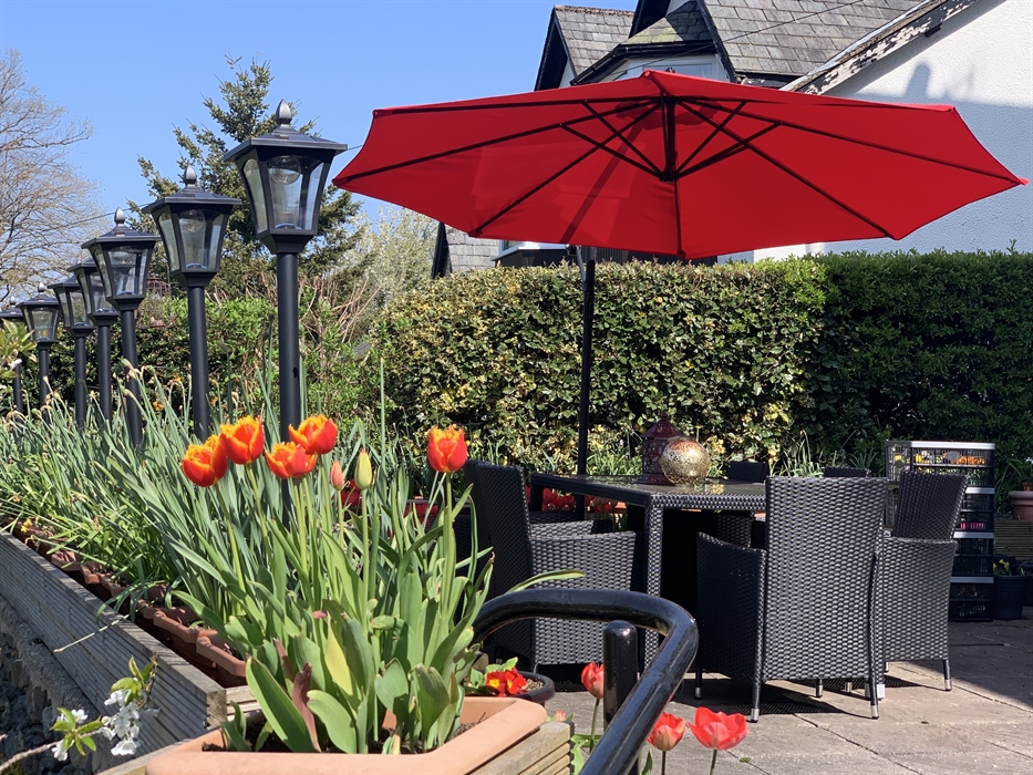 A sunny patio with a black wicker dining table and chairs beneath a large red umbrella. A row of black lamp posts lines the edge of the patio, next to