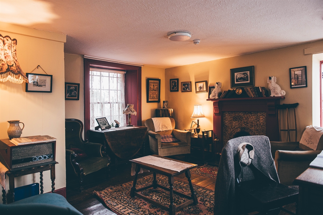 The Parlour. 1950s decorated, small room with comfy chairs and pictures on the walls. A chair with a jacket hanging over the back is positioned in fro