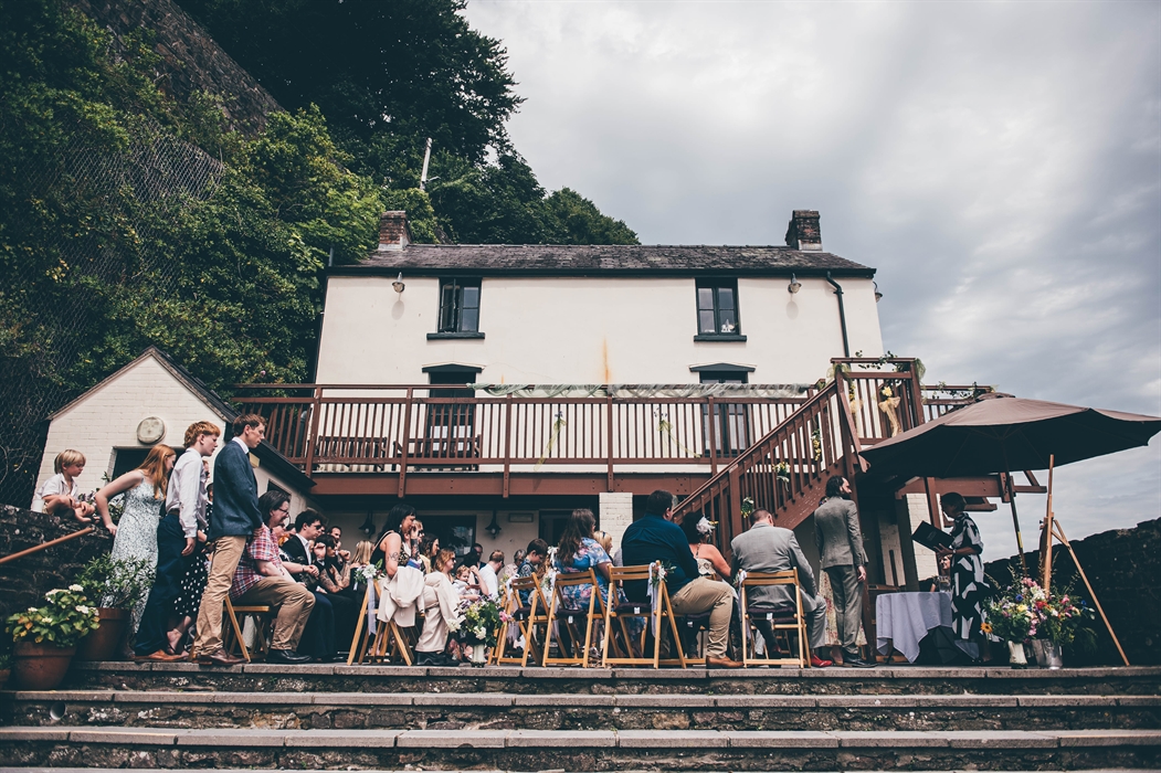 View of the back of a white-walled Boathouse looking up. A largely seated wedding party are on the patio in the foreground.