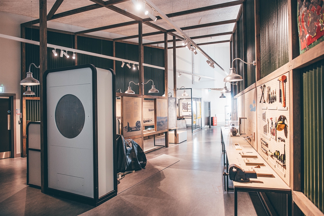 A view of a gallery space with a workbench and tools on the right and wind tunnel apparatus on the left.