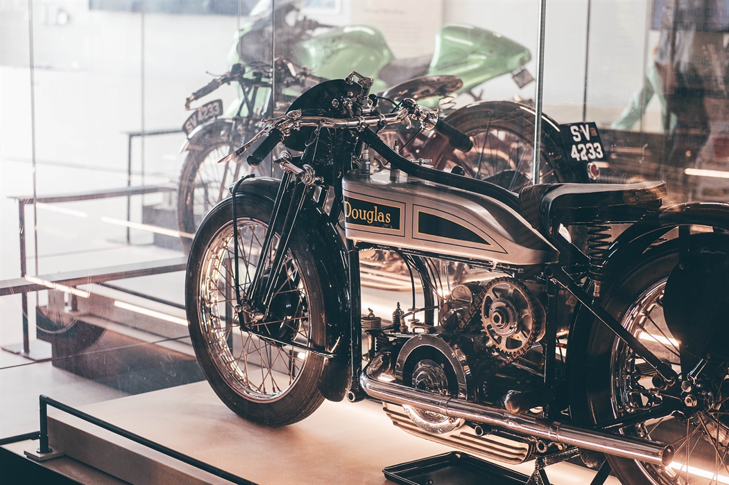 A silver Douglas motorcycle displayed in a glass case in the foreground with a green motorcycle in a case in the background