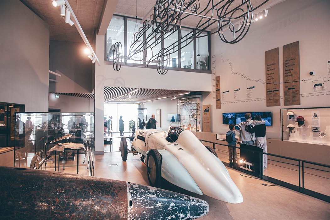 Wide angle of the main gallery interior of the museum. A large metal tail of the Bluebird car is in the foreground and the Babs car is in the middle g