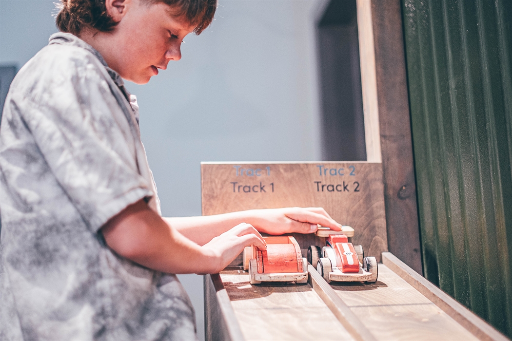 A teenage boy holds two model cars on sloped wooden tracks