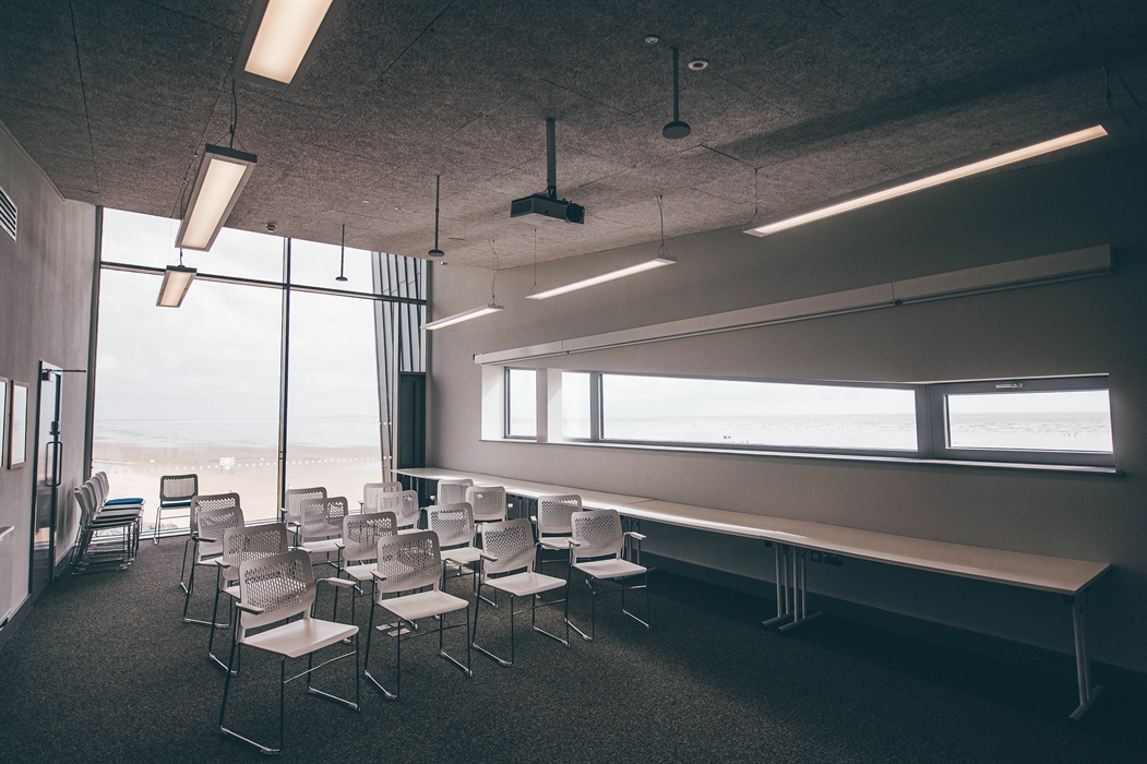 A wide angle view of a light function room. Chairs are spread in theatre style. A floor-to-ceiling window looking over a beach is in the background.