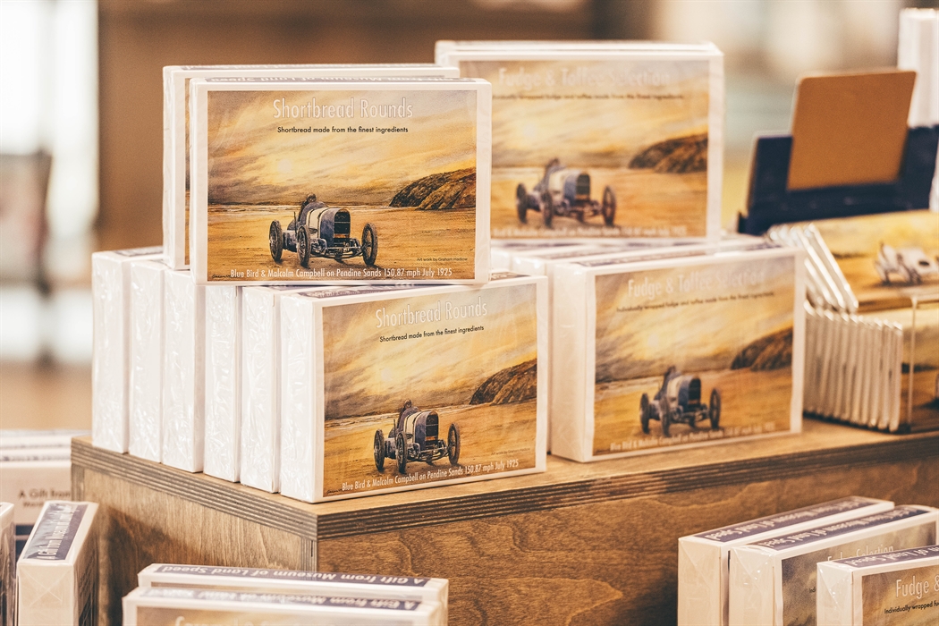A close up of a display table with boxes of fudge that have an image of a painting of the Bluebird car on Pendine Sands.