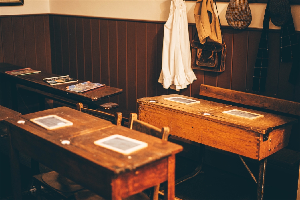 Victorian school desks with chalk writing boards on them. Victorian clothes hang on pegs in the background.