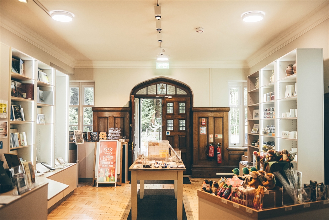 Brightly lit and well stocked gift shop with shelving units on either side and two display tables in the centre and foreground.