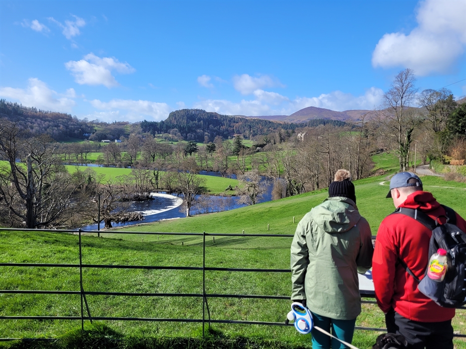 Two walkers with a dog approaching a filed overlooking a river and mountains