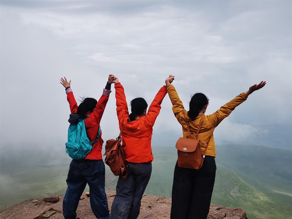Three walkers stand with their arms outstretched looking out over the mountains and clouds