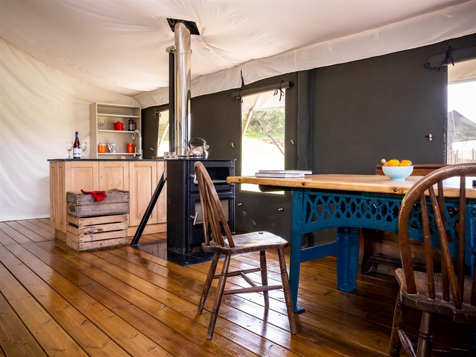 Enfys dining room, showing a big table with teal base, 2 chairs, a wood burner with oven and a slate and oak kitchen unit in the background