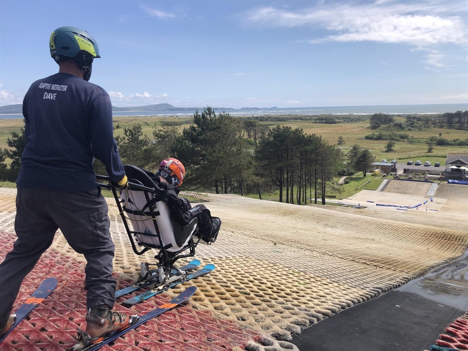 A ski instructor holds a sit ski on top of an artificial slope with views of the coastline