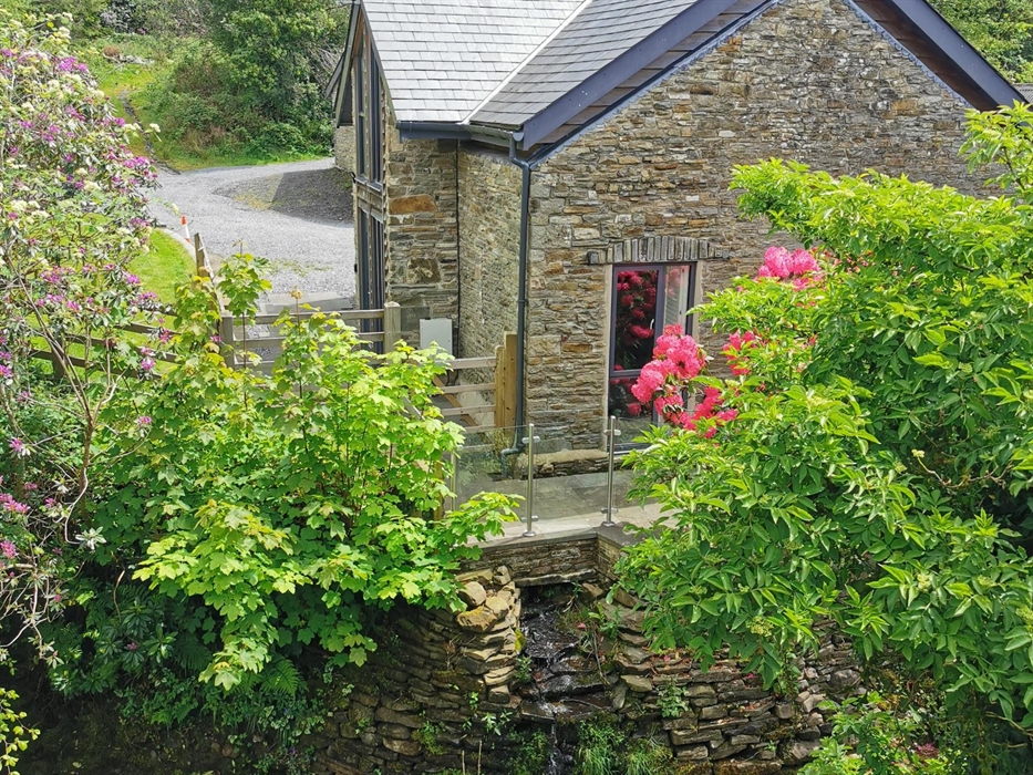 A charming stone cottage nestled among flowering shrubs and trees, with a babbling stream flowing below the patio.
