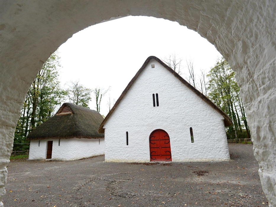 looking through a stone archway, you can see two white buildings with thatched rooves. The larger building has a big red door with black metalwork.