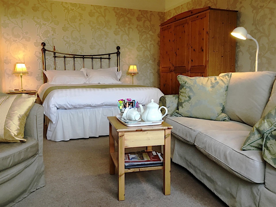 Double room showing king-size bed with traditional brass headboard, bedside lamps, sofa, and tea/coffee tray on a small coffee table