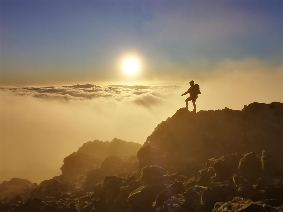 The silhouette of a walker on top of a mountain facing the sun rising above a cloud inversion.