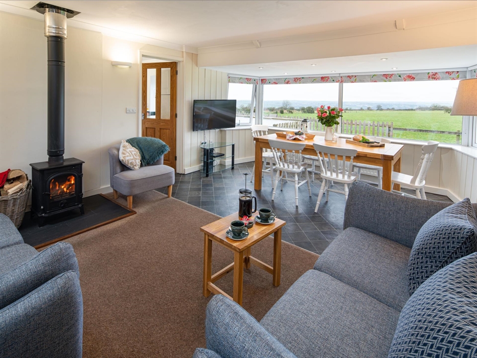 The open plan living room with countryside views of Pembrokeshire and Ceredigion.  Comfy sofas, glowing log burner and coffee table in the foreground,