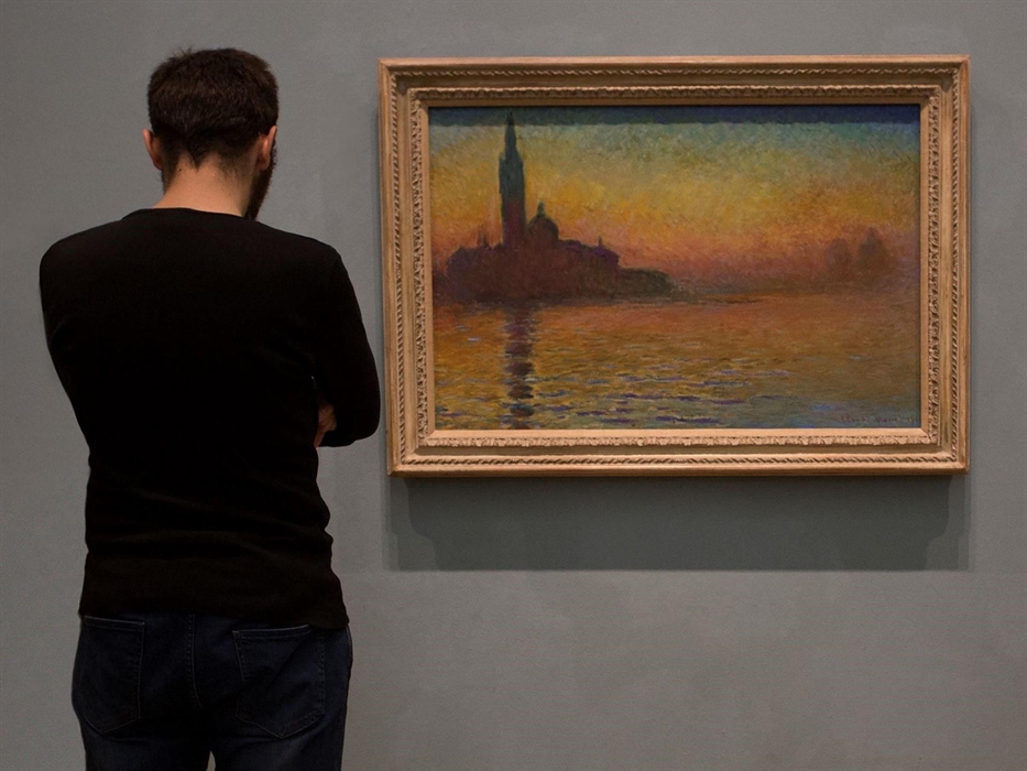 Young man with his back to the camera. He's looking at a painting showing the silhouette of a church. the sky and the reflection of the church in the