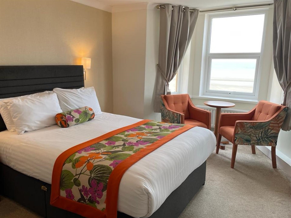 One of our superior double rooms, picture showing a King size bed with orange floral runner, 2 armchairs and a side table in the window overlooking th