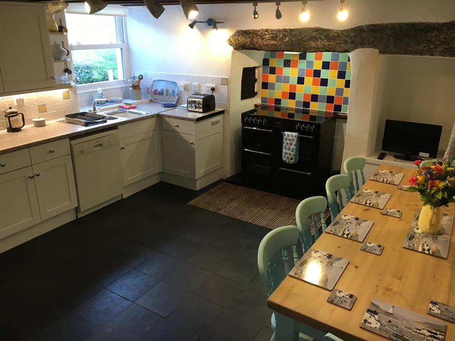 Kitchen at Talwrn Bach Farmhouse, shows modern kitchen units, a country table and chairs, range cooker and ancient stone lintel